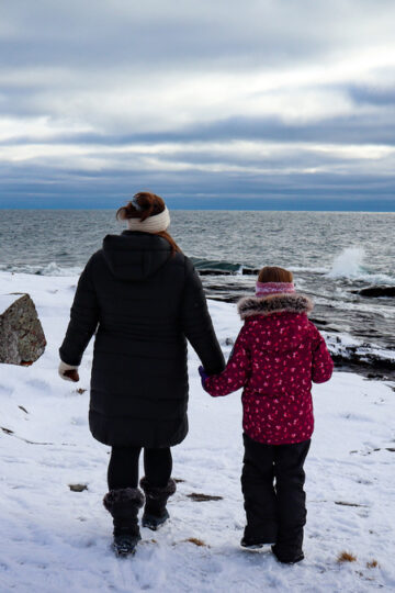A mom and her young daughter walk along the snow toward Lake Superior in their winter gear.
