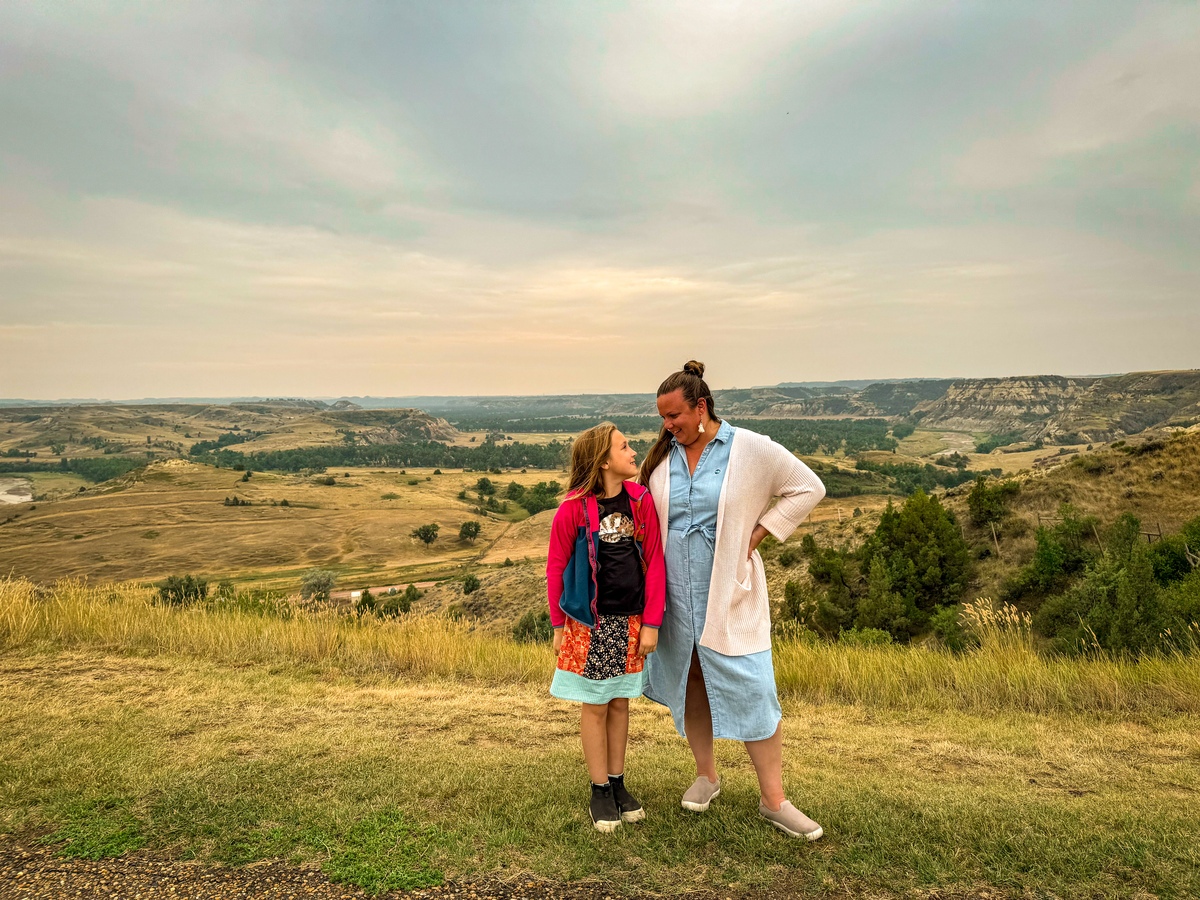 A woman and her young daughter stand together with a view of the North Dakota badlands in the distance.