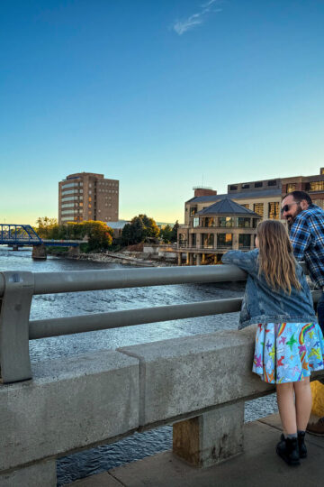 A father and daughter stand together on a bridge looking at the Grand River.